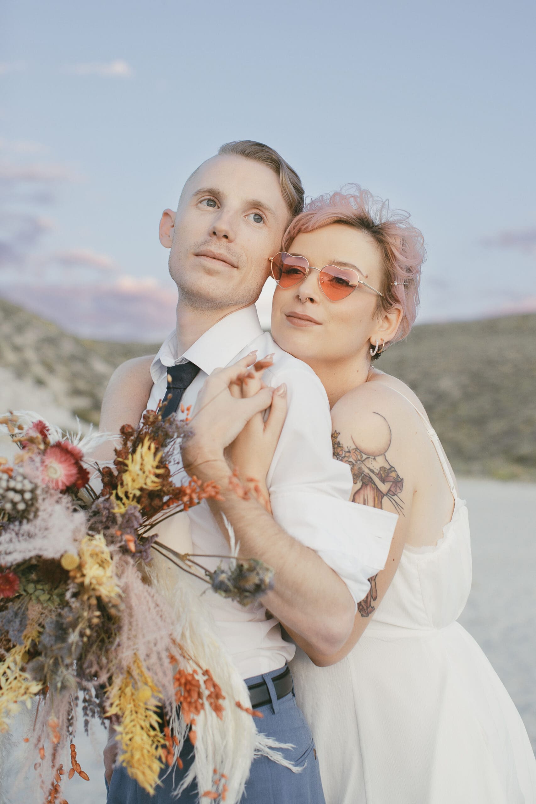 A bride wearing wearing heart shaped glasses hugs her groom from behind while they look off into the distance at a sunset.