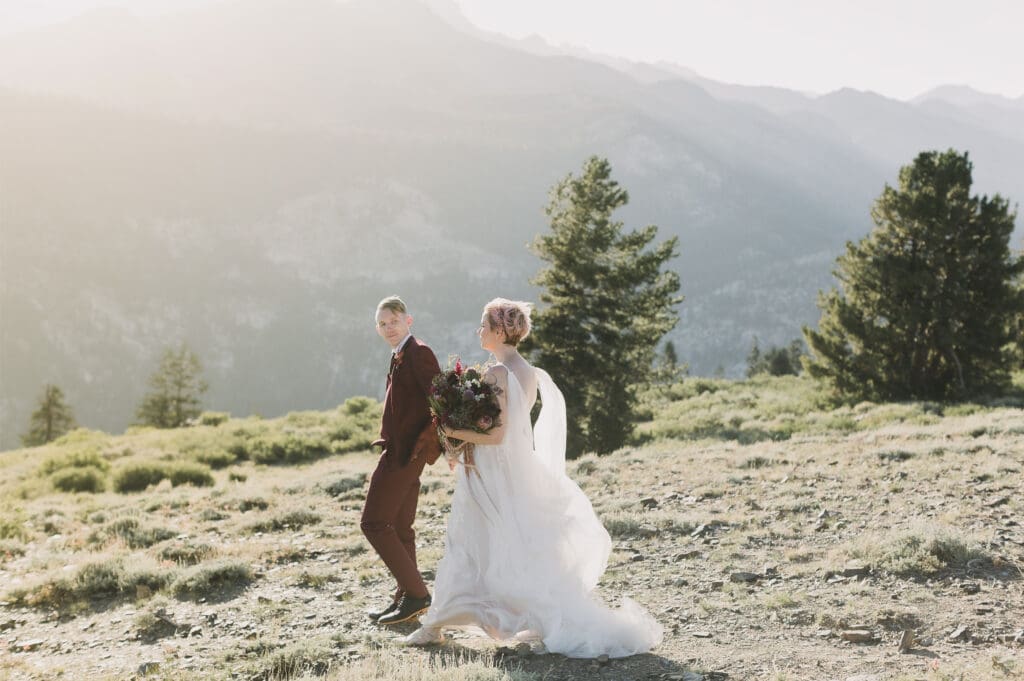 A groom leads the way down a mountain side as a bride follows, while in view of a giant mountain in Mammoth Lakes, California. 