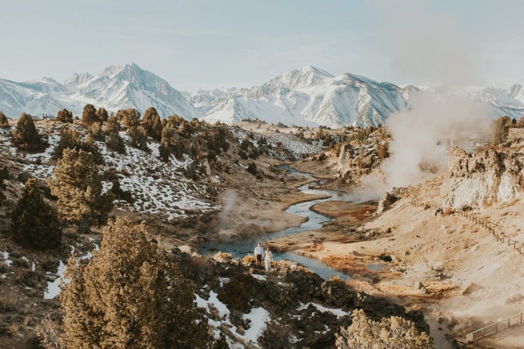 A bride and groom stand in front a snow covered mountains near at Hot Creek Geological site near Mammoth Lakes, California.
