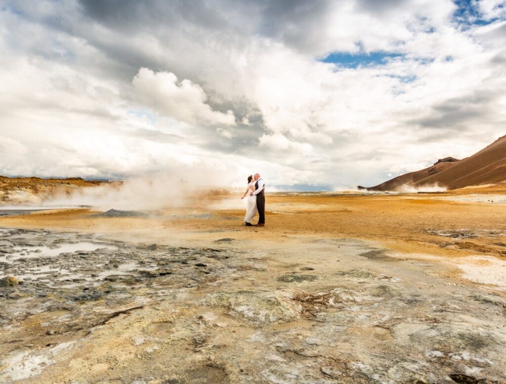 A bride and groom stand in a surreal, alien looking background of organse sand, bright clouds and steam in Iceland.