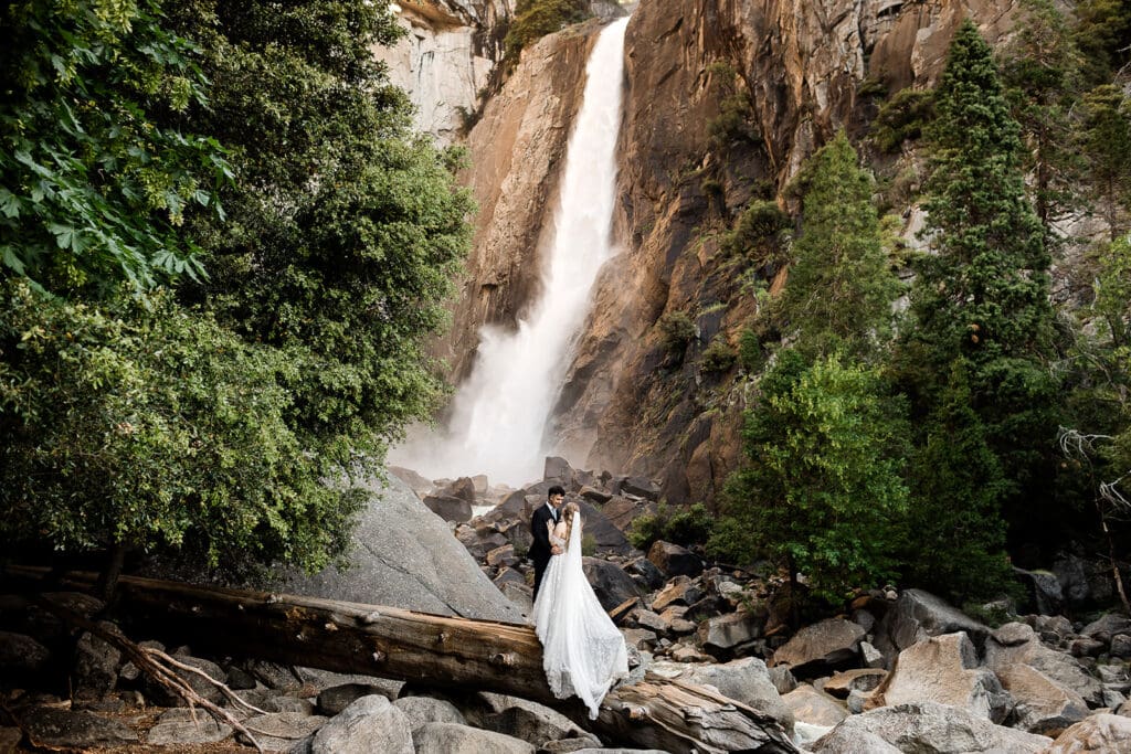 A bride and groom stand in front of lower yosemite falls on their wedding day.