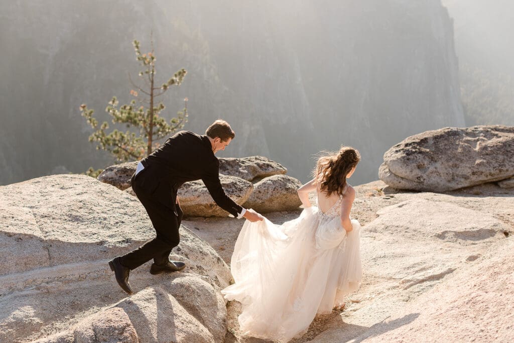 A groom helps his bride as she jumps down a rock at Taft Point