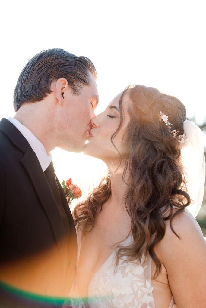 A bride and groom kiss in bright sunlight.
