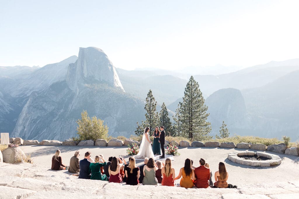 A small group of guests watch while a bride adnd groom say their vows in front of Half Dome in Yosemite.