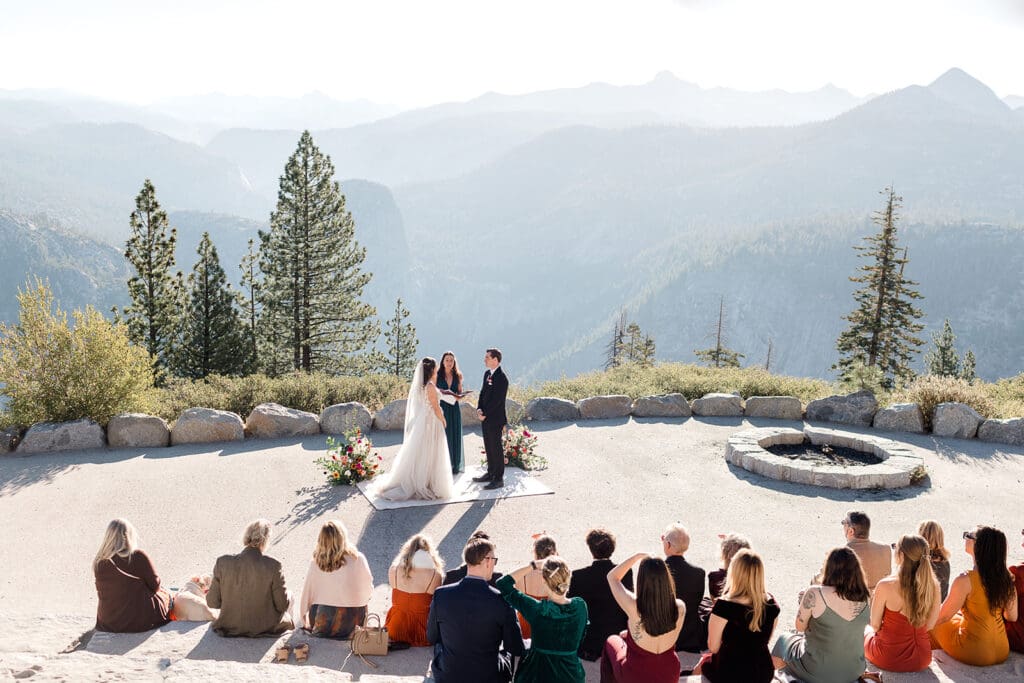A small group of guests watch while a bride adnd groom say their vows in front of Half Dome in Yosemite.