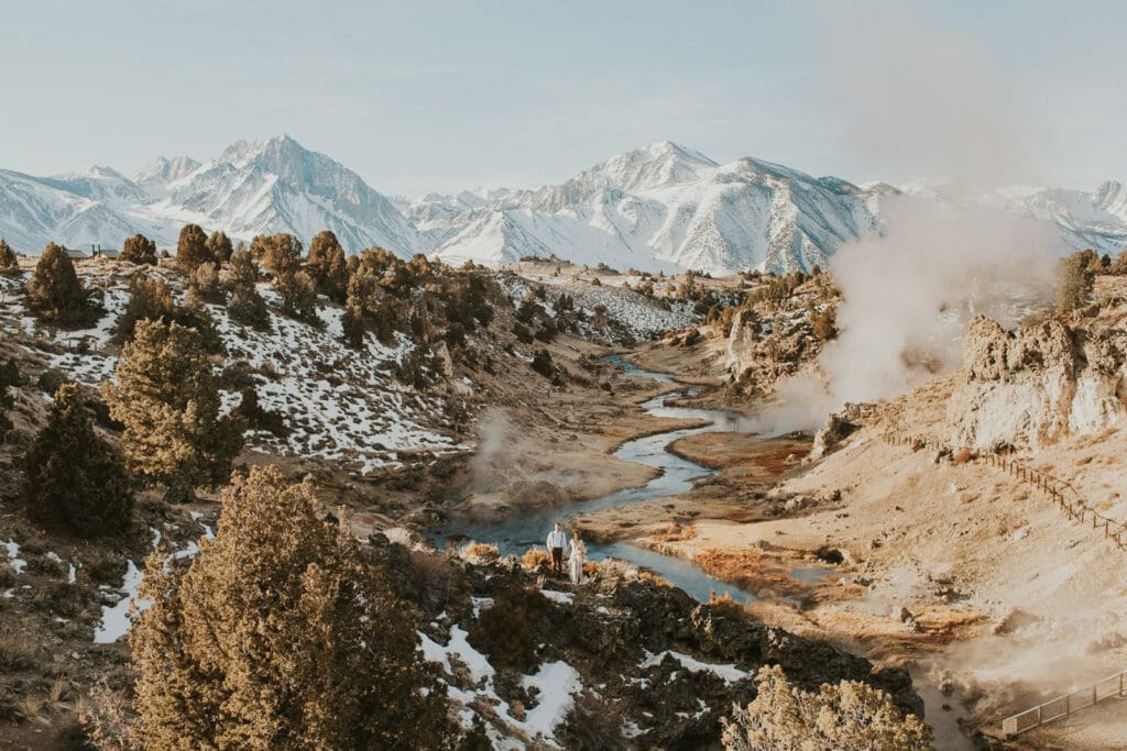 A bride and groom look towards snow covered mountains and steamy hot creek on their wedding day in Mammoth Lakes.